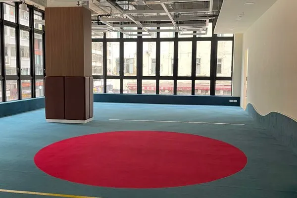Ref#3216: A spacious, modern office rental with large windows, blue carpet, and a bold red circle centerpiece. Features exposed ceiling pipes, a wood-paneled column for style, and abundant natural light throughout the space.
