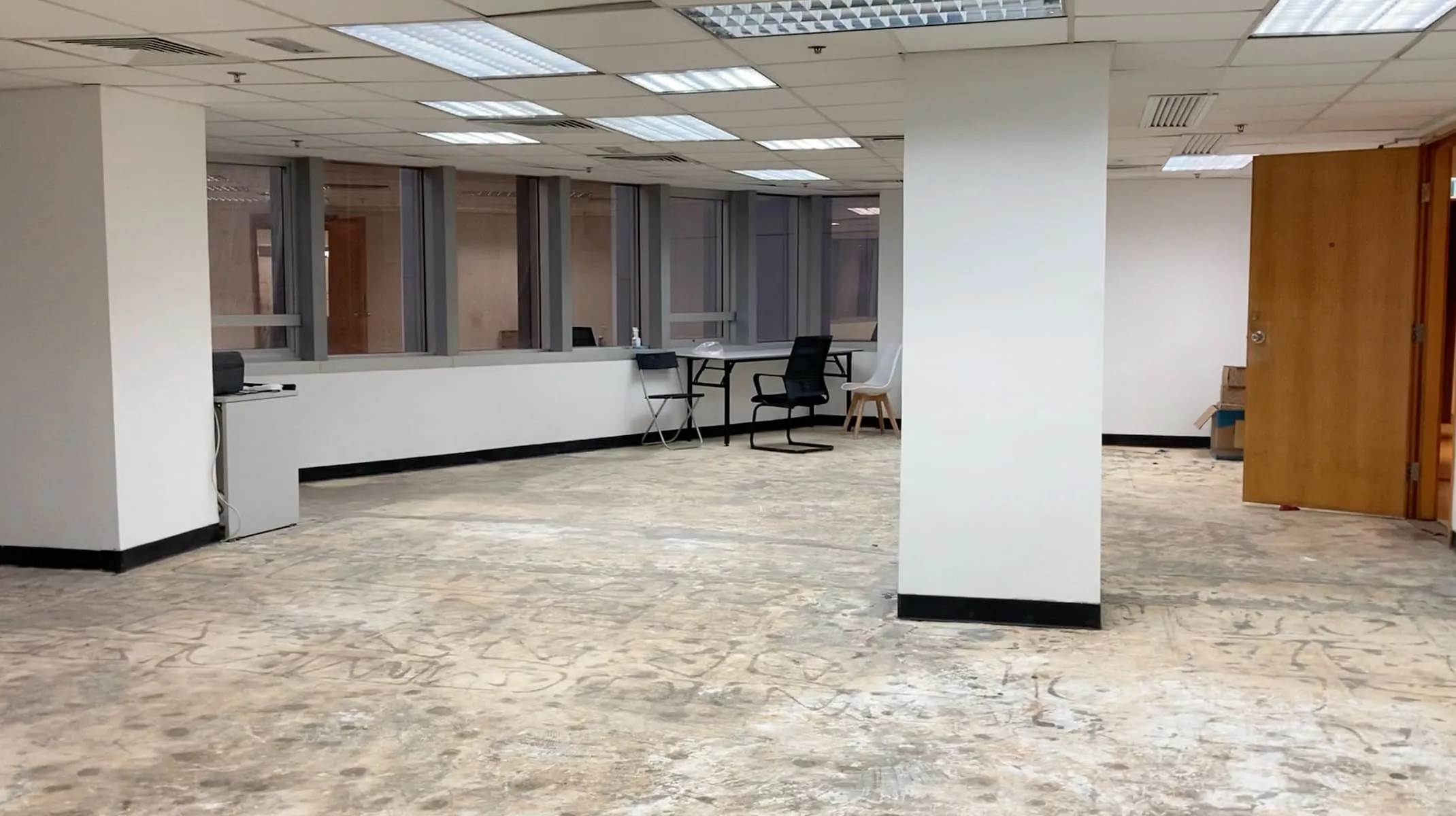 Ref#3224 is an empty office space for rent, offering exposed concrete floors, a white central pillar, large back windows, fluorescent ceiling lights, and several scattered chairs and desks.
