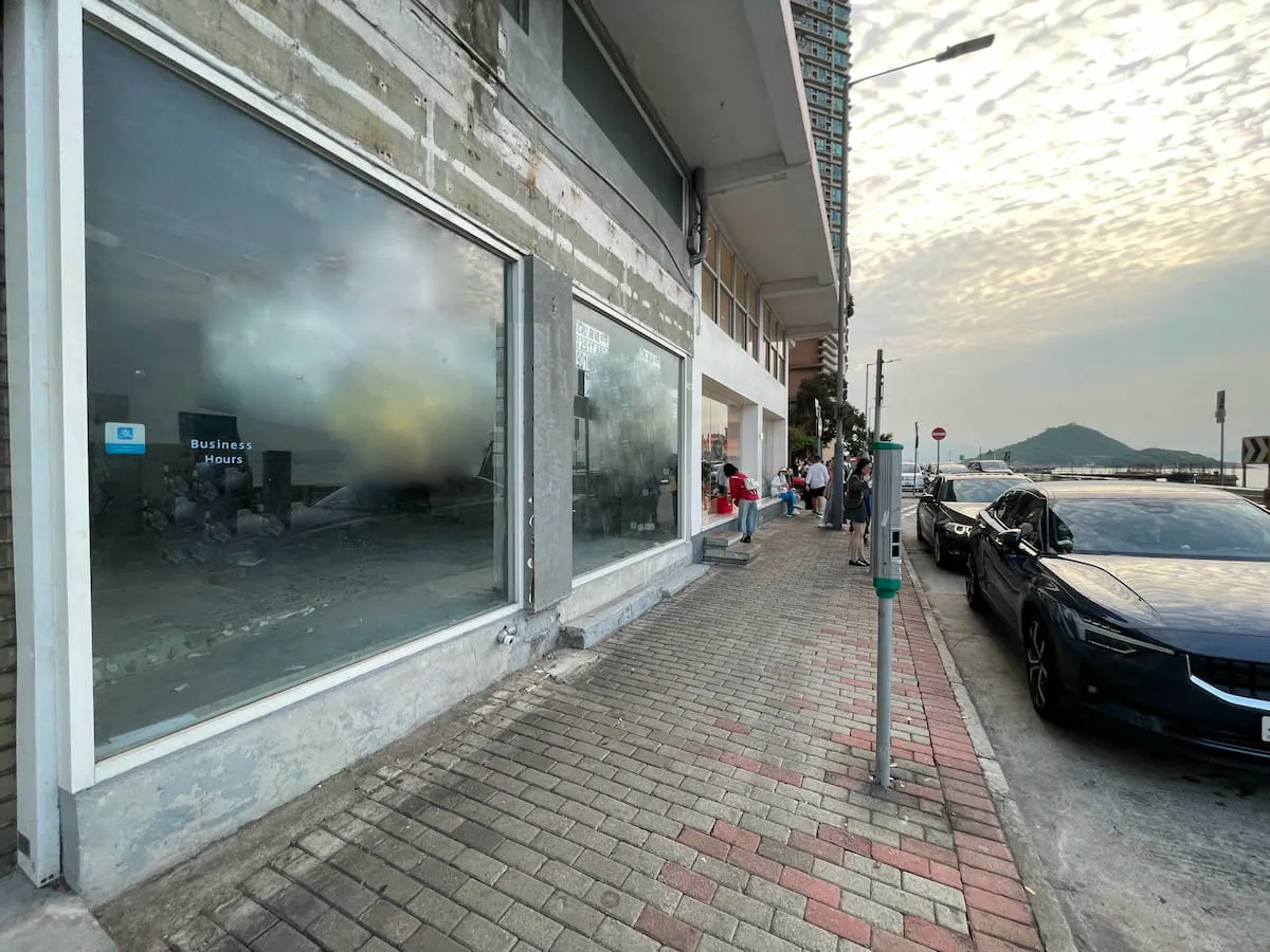 Ref#3608: A sidewalk runs along the building9s large fogged-up windows	68perfect for shop rentals. People gather at the entrance, cars line the curb, and a cloudy sky frames the hill in the background.