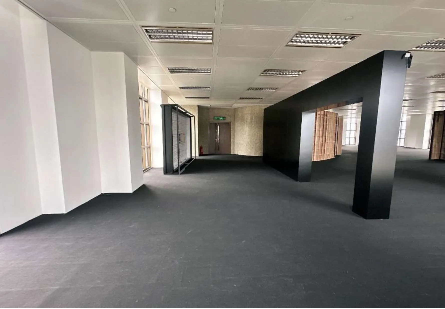 Ref#3609 features a spacious room with black carpet, white walls, ceiling lights, an emergency exit sign over a closed door, and large windows along one side—ideal for renting as an office or shop.