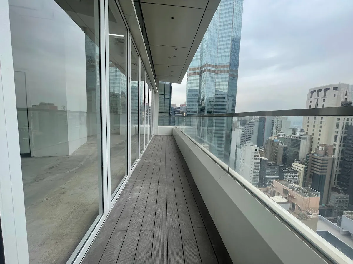 Ref#3616 features a modern narrow balcony with wooden flooring, glass railings, and floor-to-ceiling windows overlooking city skyscrapers ideal for renting office space in the city center on a cloudy day.
