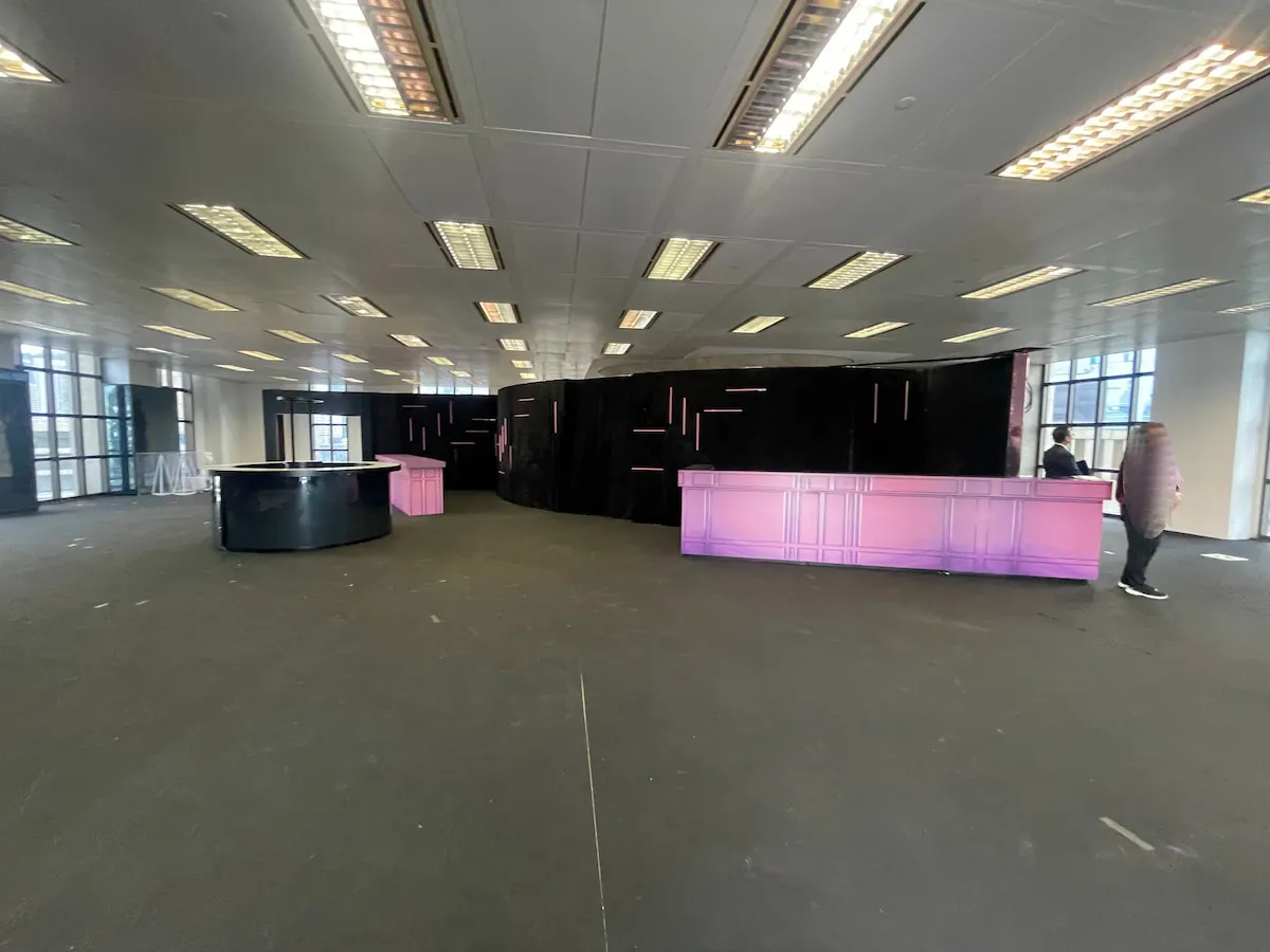 Ref#3609: Spacious modern room with high ceiling, fluorescent lights, and large windows—perfect for shop rental. Features a curved, purple-lit counter on the right, a round black counter on the left, and two people by the counters.