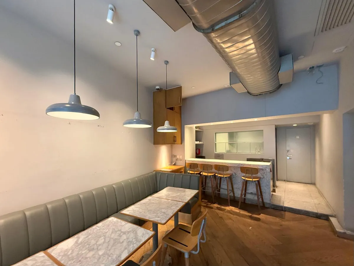 Ref#3636 is a modern, minimalist café interior for rent, featuring marble tables, wooden chairs, a padded bench, pendant lights, a marble bar counter, and an exposed air duct. The light neutral palette gives an airy vibe—perfect for a shop or restaurant.