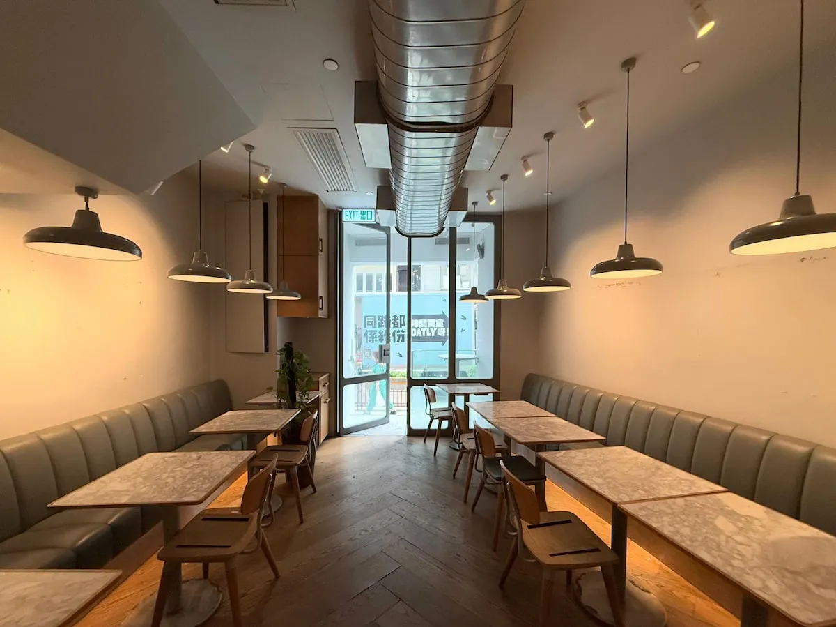 Ref#3636: A modern, empty cafe with marble tables, wooden chairs, green cushioned bench seating, and pendant lights—ideal for restaurant rental. Large windows at the back let in natural light; two people are visible outside through the glass door.
