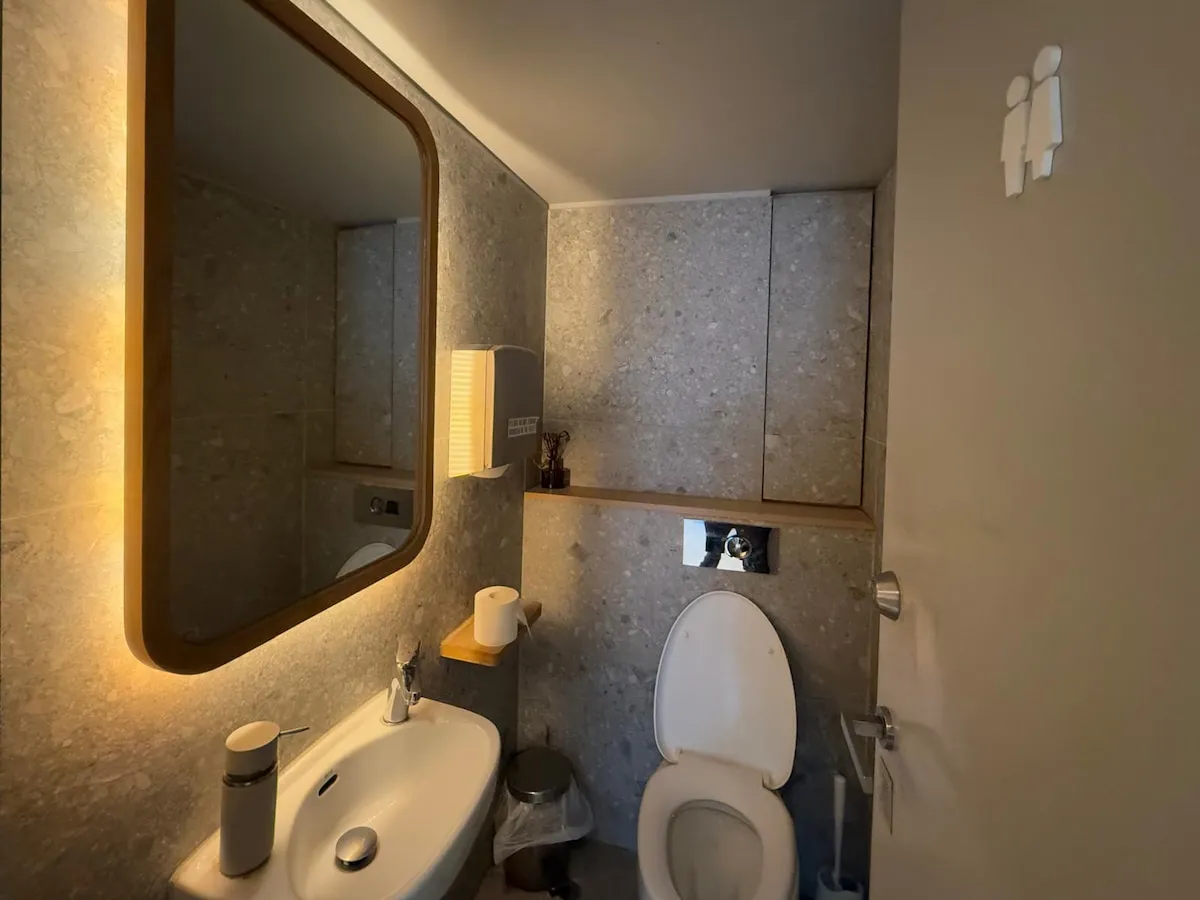 Ref#3636: A compact modern bathroom ideal for restaurant or office rentals, featuring a lighted mirror, sink, soap dispenser, toilet (seat up), toilet paper, trash bin, and a wall-mounted unisex restroom sign.