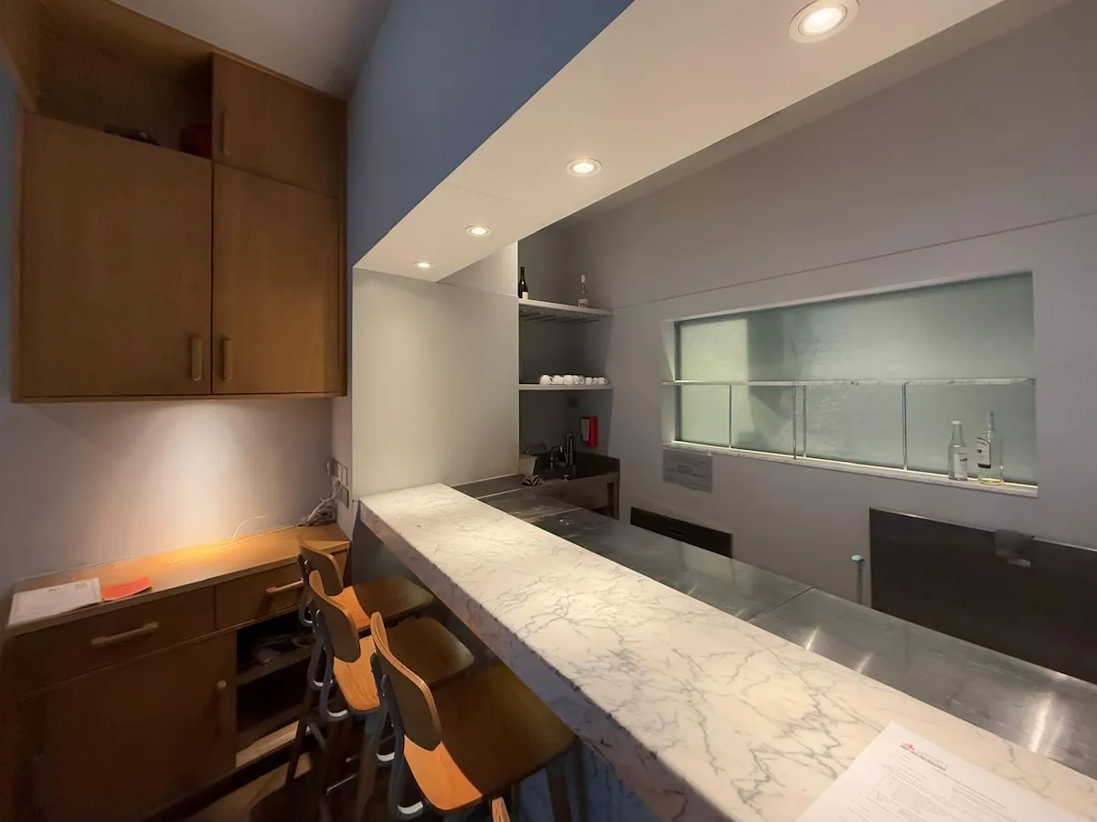 Ref#3636 offers a modern minimalist kitchen with marble countertop, three wooden barstools, light wood cabinets, recessed lighting, and a compact workspace—perfect for stylish restaurant rental or shop space.
