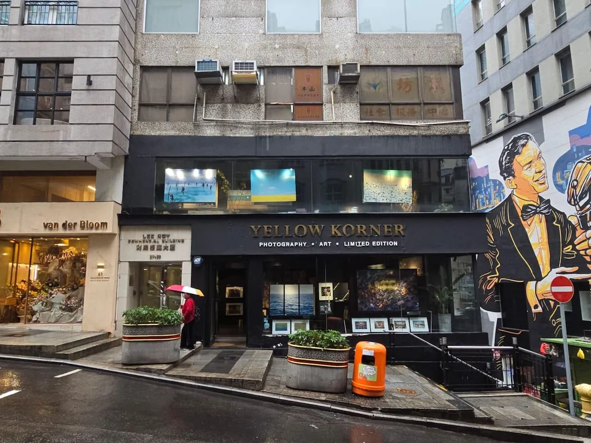 雨天下Yellow Korner畫廊街景,櫥窗展示藝術品,一人撐粉紅色雨傘走過。
