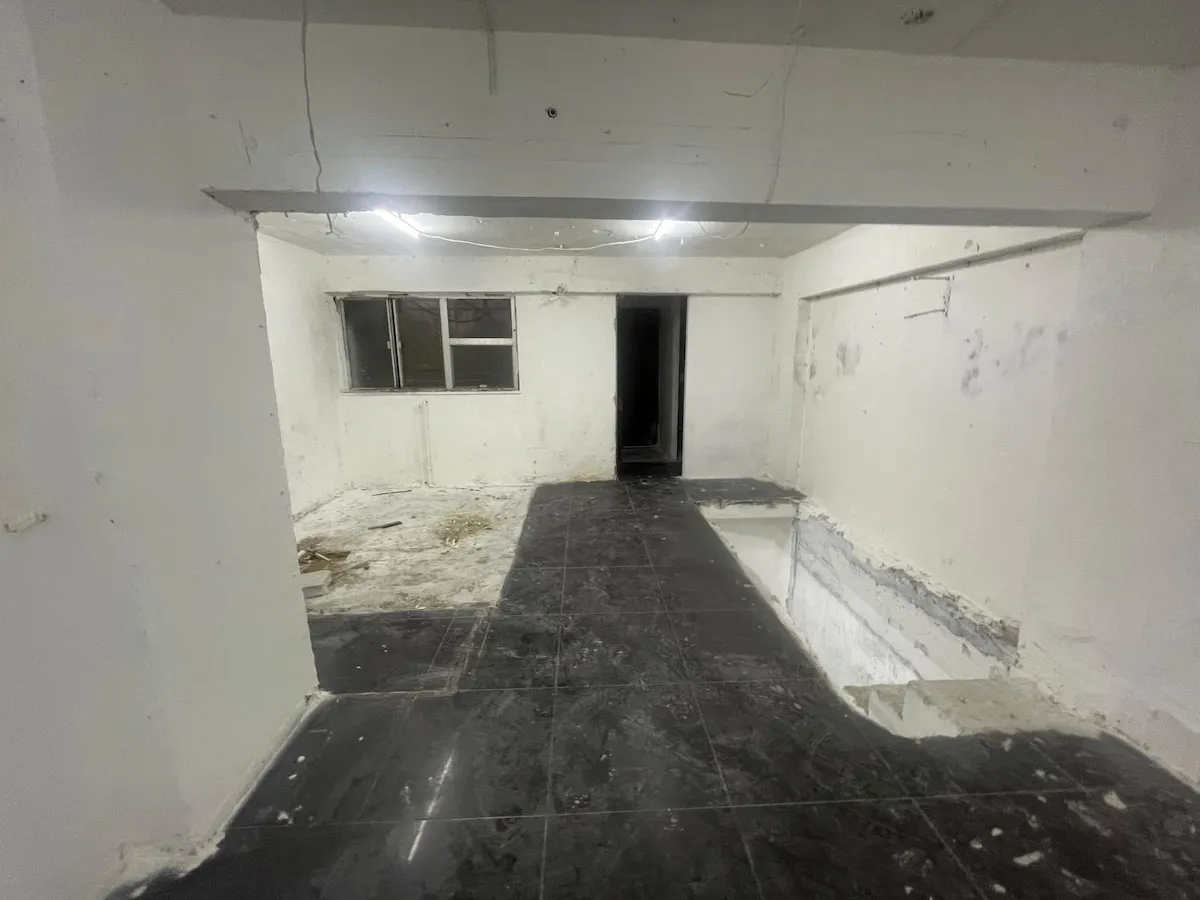Ref#3623: An empty, unfinished room with dirty white walls and black tiled floor, ideal to rent as a shop or office. Features an open window, dark doorway, and areas under renovation with exposed walls and debris.