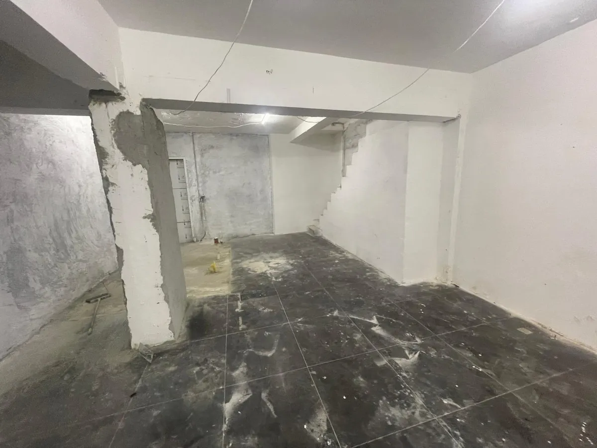 Ref#3623 features a bare, unfinished basement with rough concrete floors, unpainted white walls, exposed wires, a partial staircase, and construction debrisideal to rent as shop or office space.