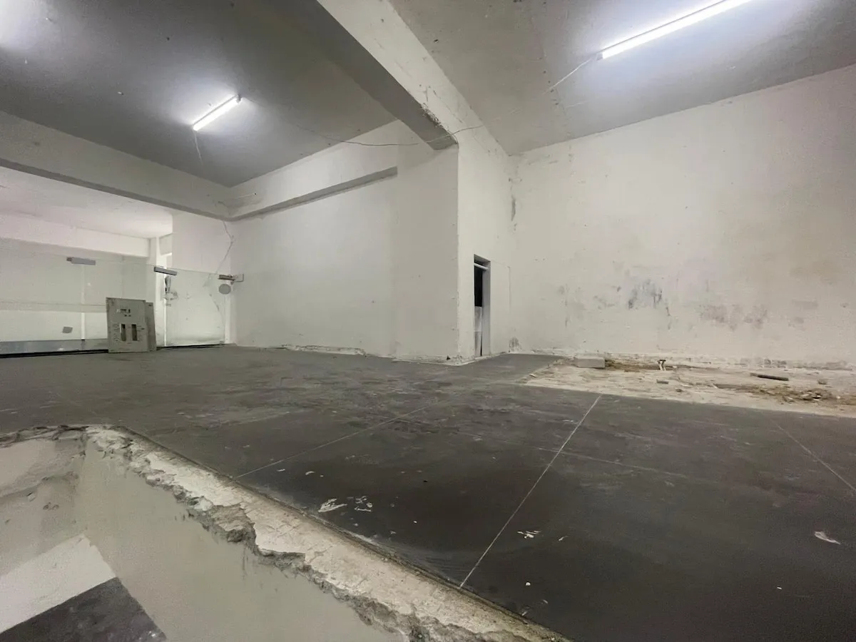Ref#3623: Unfinished indoor space with white walls, exposed ceiling, tiled but uneven/damaged floor, and fluorescent lightsideal to rent as a restaurant or shop.