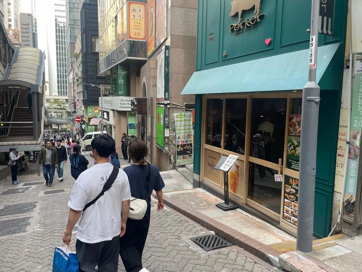 People walk along a sloped city street lined with shops and restaurants, some advertising rent opportunities. A green awning covers a restaurant entrance on the right. Product Name: Ref#3623.
