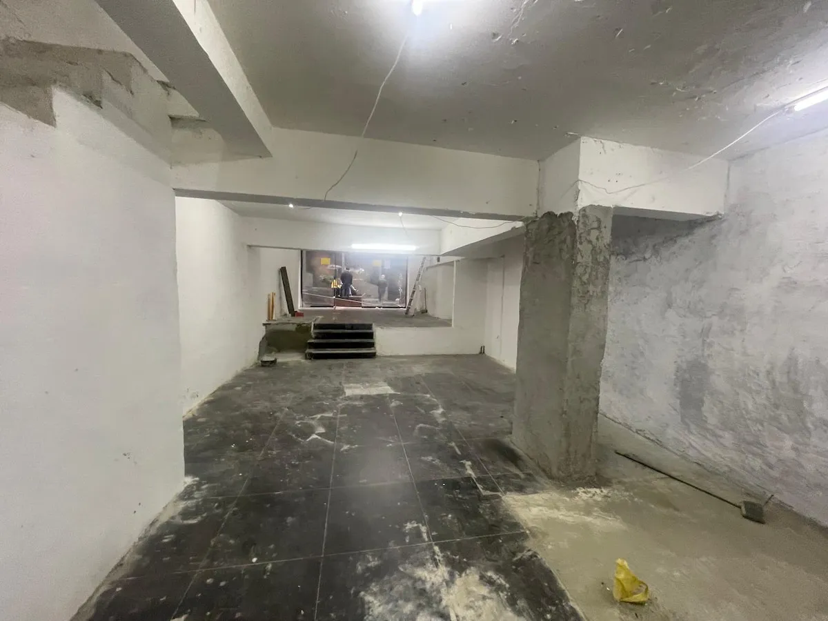Ref#3623: Partially renovated basement with white walls, black unfinished tile flooring, scattered construction materials, and bright overhead lightingideal to rent as a shop or office. Stairs lead up to workers in the distance.