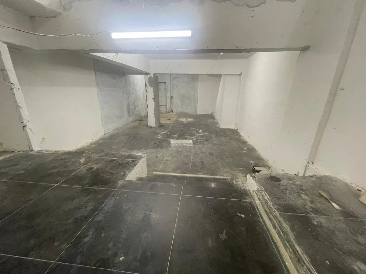 Ref#3623: A dimly lit, empty basement with unfinished concrete walls and black tiled floorssome tiles missing or damaged. Low ceiling with exposed pipes and wires offers an industrial vibe, ideal for office or shop rental.