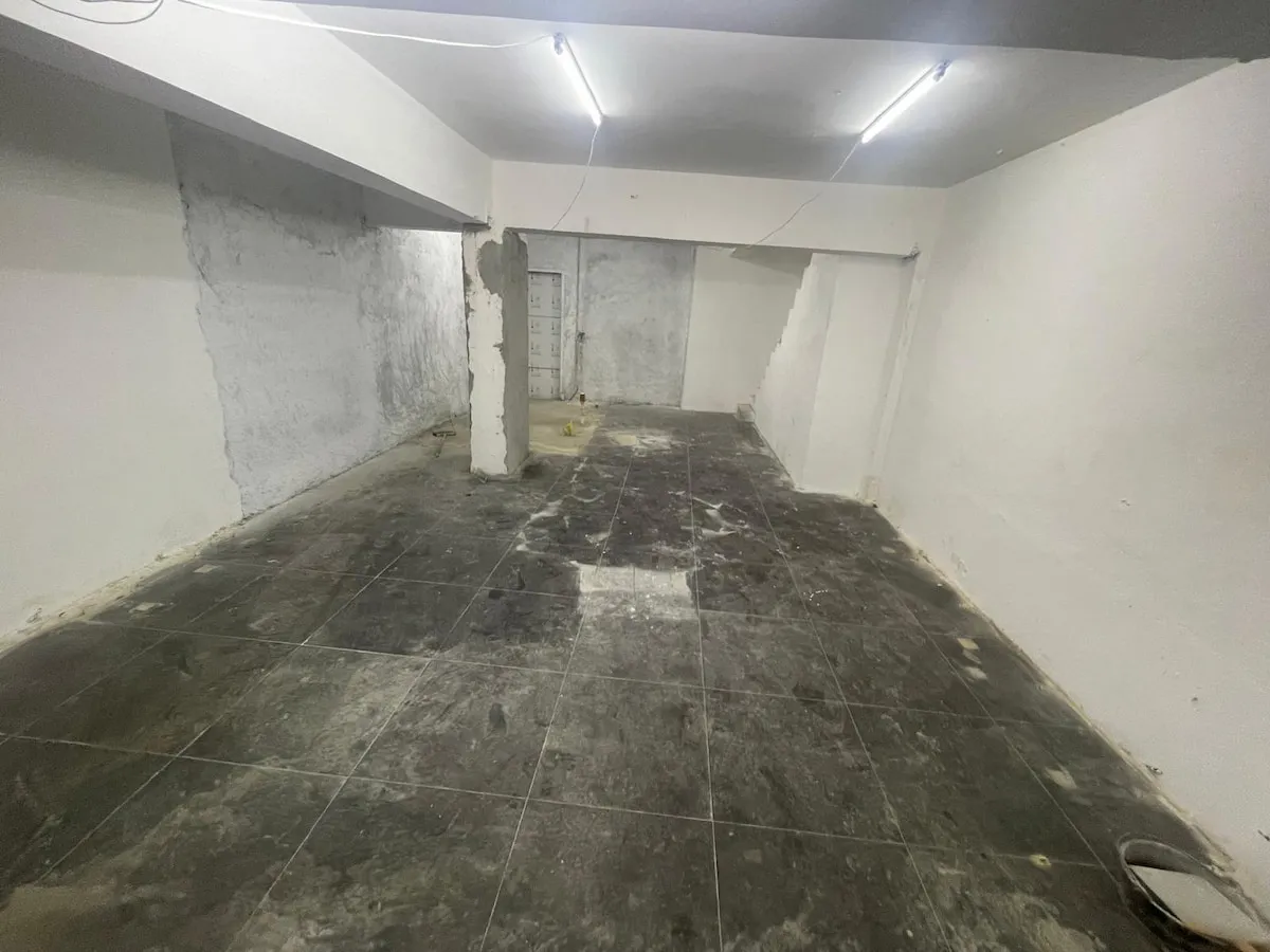 Ref#3623: An empty, dimly lit room with unfinished white walls, dark tiled floor, exposed wires from the ceiling, scattered debris, and a closed doorideal for rent as a shop or office.