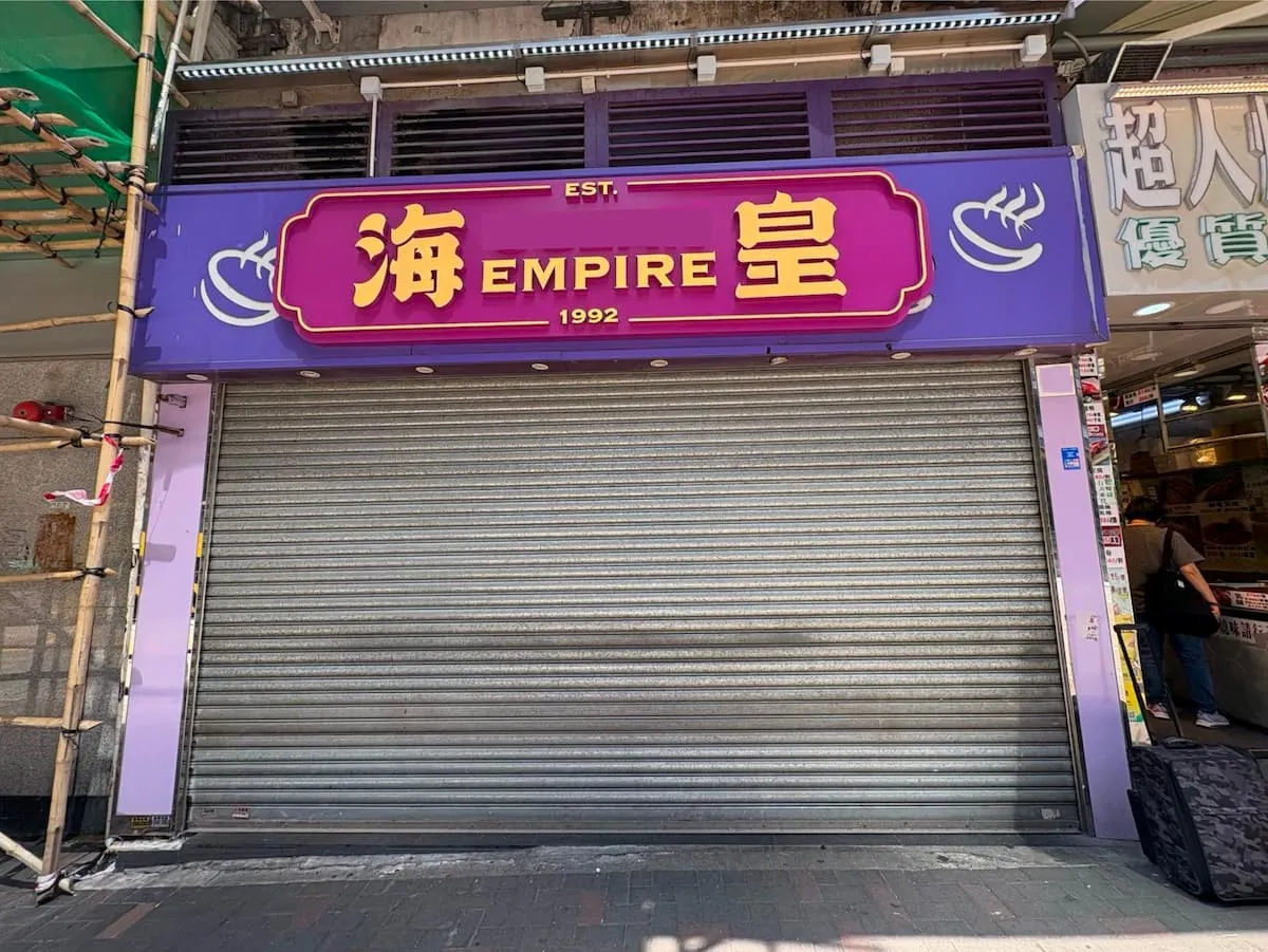 Ref#3625: A storefront with a purple EMPIRE sign in English and Chinese (est. 1992). The closed metal shutter hints at possible shop space for rent. Sidewalk and nearby stores are also visible.