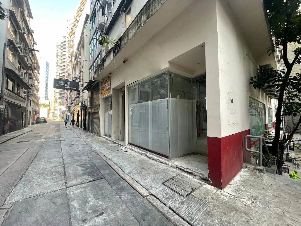 Ref#3626: A narrow urban street with old buildings and a quiet, slightly worn feel. Features a corner shop with glass windows covered by white sheetsideal for shop rental. Two people are seen walking away in the distance.
