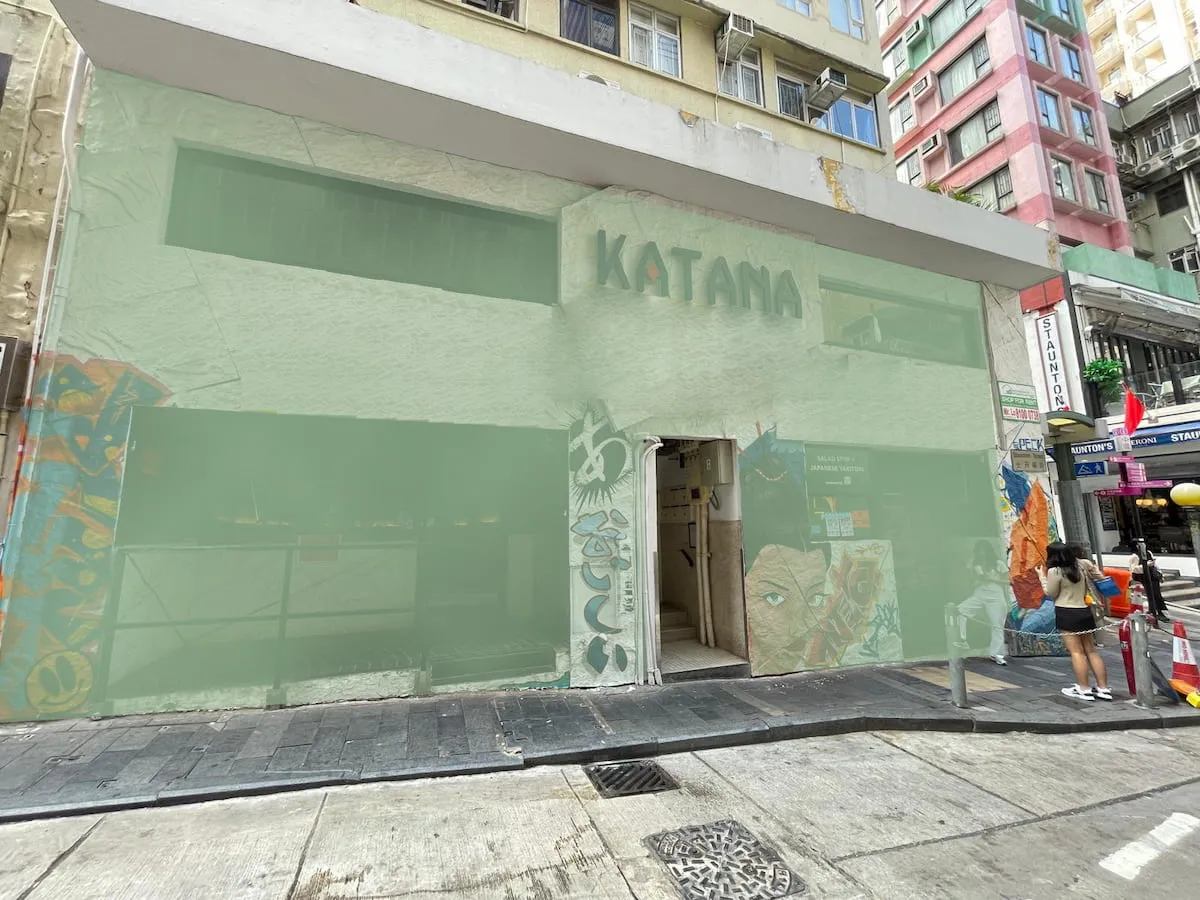 Street view of building Ref#3627 with "KATANA" signage, wall art by the door, and facade wrapped in green construction nettingperfect for shop rental; person visible on sidewalk.
