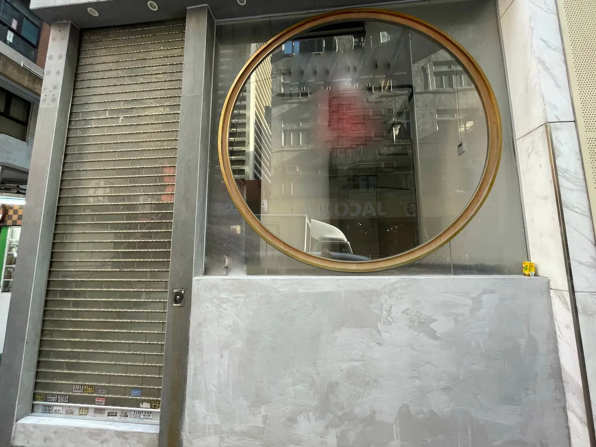 Ref#3633 is a shop rental storefront with a large gold-framed circular window, closed metal shutter on the left, and marble-like gray walls. Reflections of nearby buildings and the street can be seen in the window.