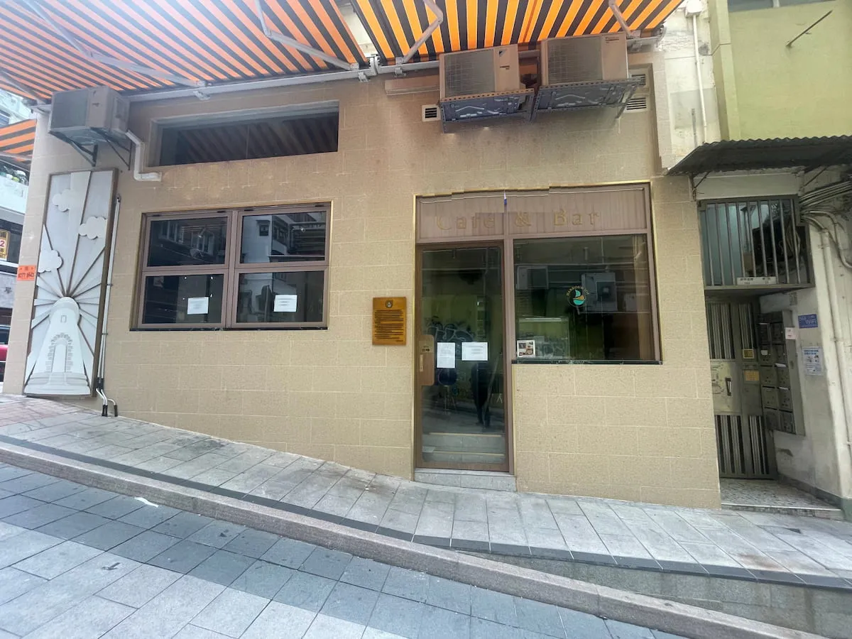 Ref#3634: Beige building with glass door/windows, orange-and-black striped awnings, and AC units on a sloped, tiled streetideal for renting as shop or office space. Notices displayed in front.