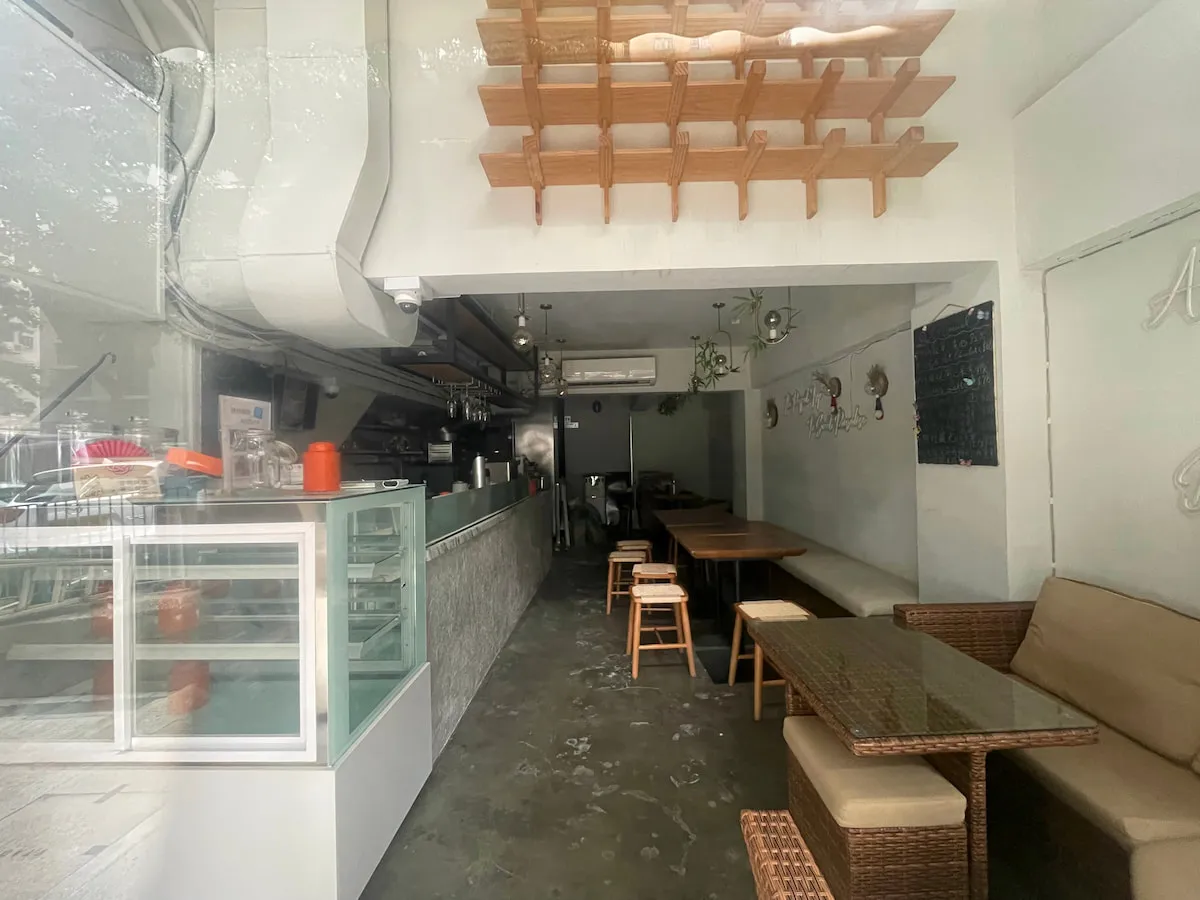 Ref#3637: Small modern café for rent featuring glass display case, wicker seats, wooden tables, white walls, wall art, ceiling shelves. Minimalist interior; well-lit and empty—great opportunity for a new shop.