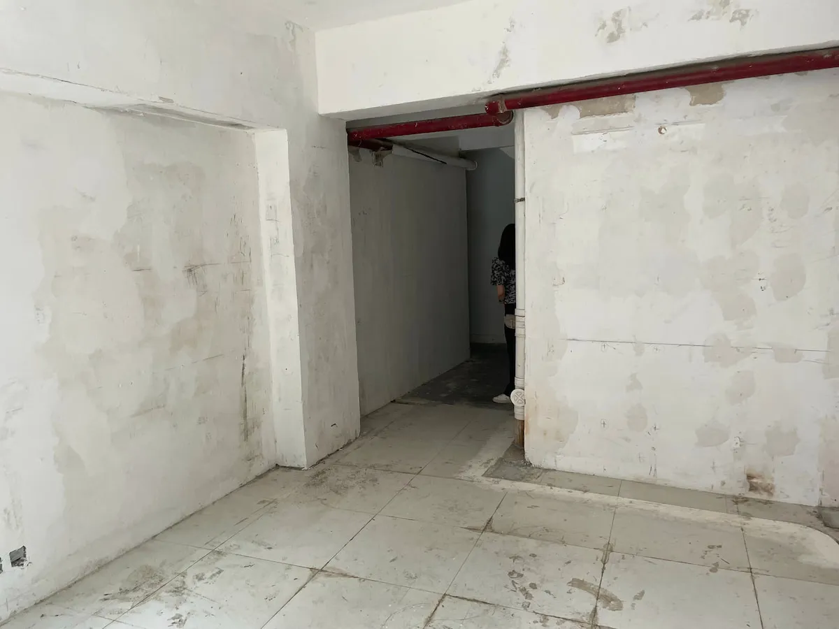Ref#3638: Spacious, unfinished room with tiled floors and worn white walls, suitable for use as a shop or office. Features red ceiling pipes and a shadowy back doorway. The space shows signs of neglect and ongoing renovation.