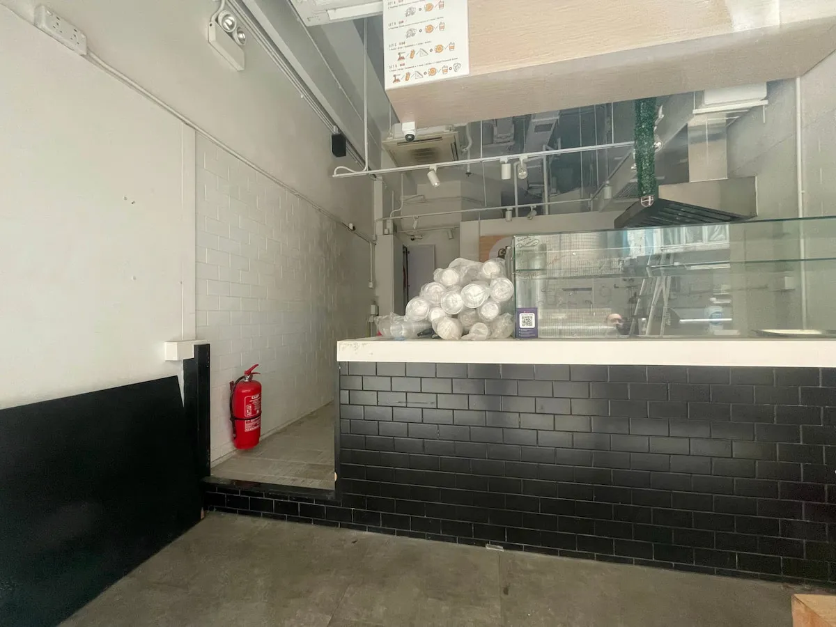 Ref#3638: A small, empty restaurant for rent featuring a black-tiled counter with plastic-wrapped items, a fire extinguisher on the left, and white tiled walls with ceiling pipes in the background.