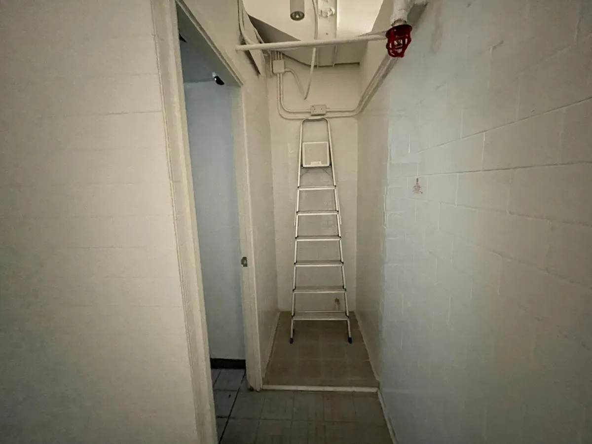 Ref#3638 features a narrow, dimly lit hallway with white brick walls, a metal ladder beneath exposed pipes and a red sprinkler0ideal as a unique rent office or restaurant rental space. An open doorway leads to a tiled room on the left.