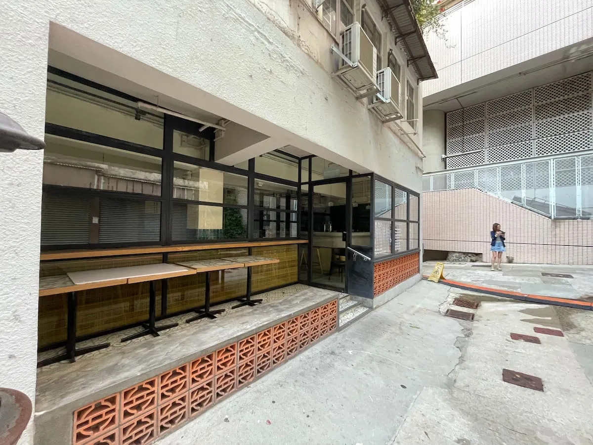 Ref#3640: A small, empty cafe with glass walls and tables inside, set in a concrete alleyideal for shop rental. A person stands near a staircase by a building with a metal grid facade.