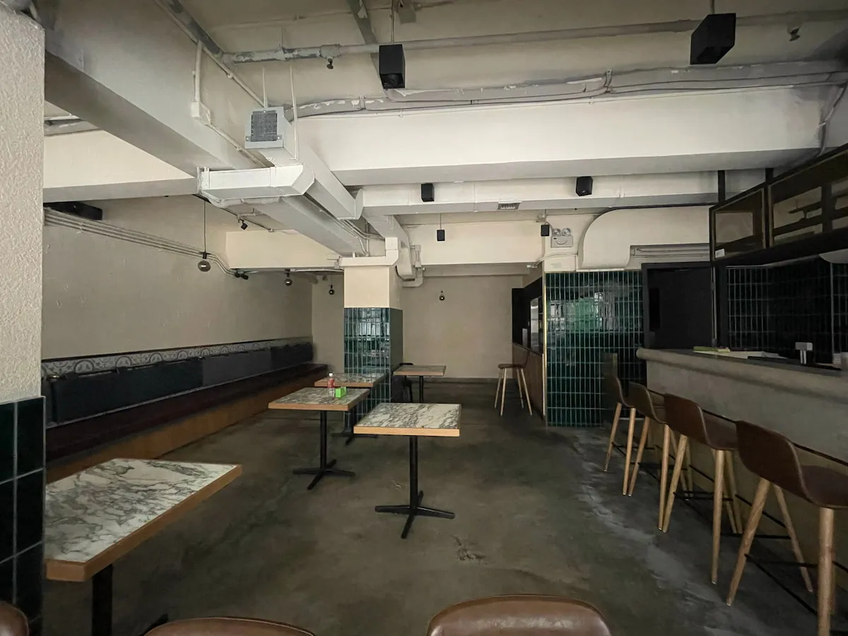 Ref#3640: A dimly lit, empty cafe with marble tables, wooden chairs, a green tiled bar, and concrete floors. Features exposed pipes, overhead lighting, left wall bench seatingideal to rent as an office or restaurant.