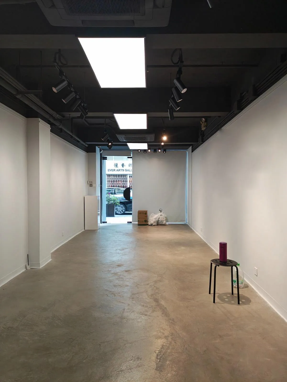 Ref#3645: Mostly empty art gallery with white walls and concrete floor. Trash bags and boxes near entrance suggest potential for office or shop rental. Small round table with water bottle and tumbler on the right.