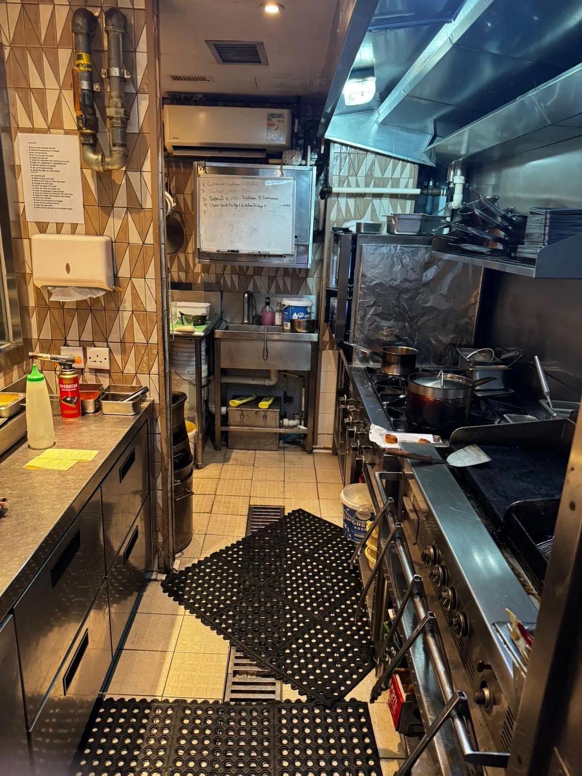 Ref#3654 is a compact commercial kitchen available for rent, featuring stainless steel appliances, stove cookware, a wall whiteboard, patterned tiles, black rubber floor mats, and counters stocked with utensils and ingredients.