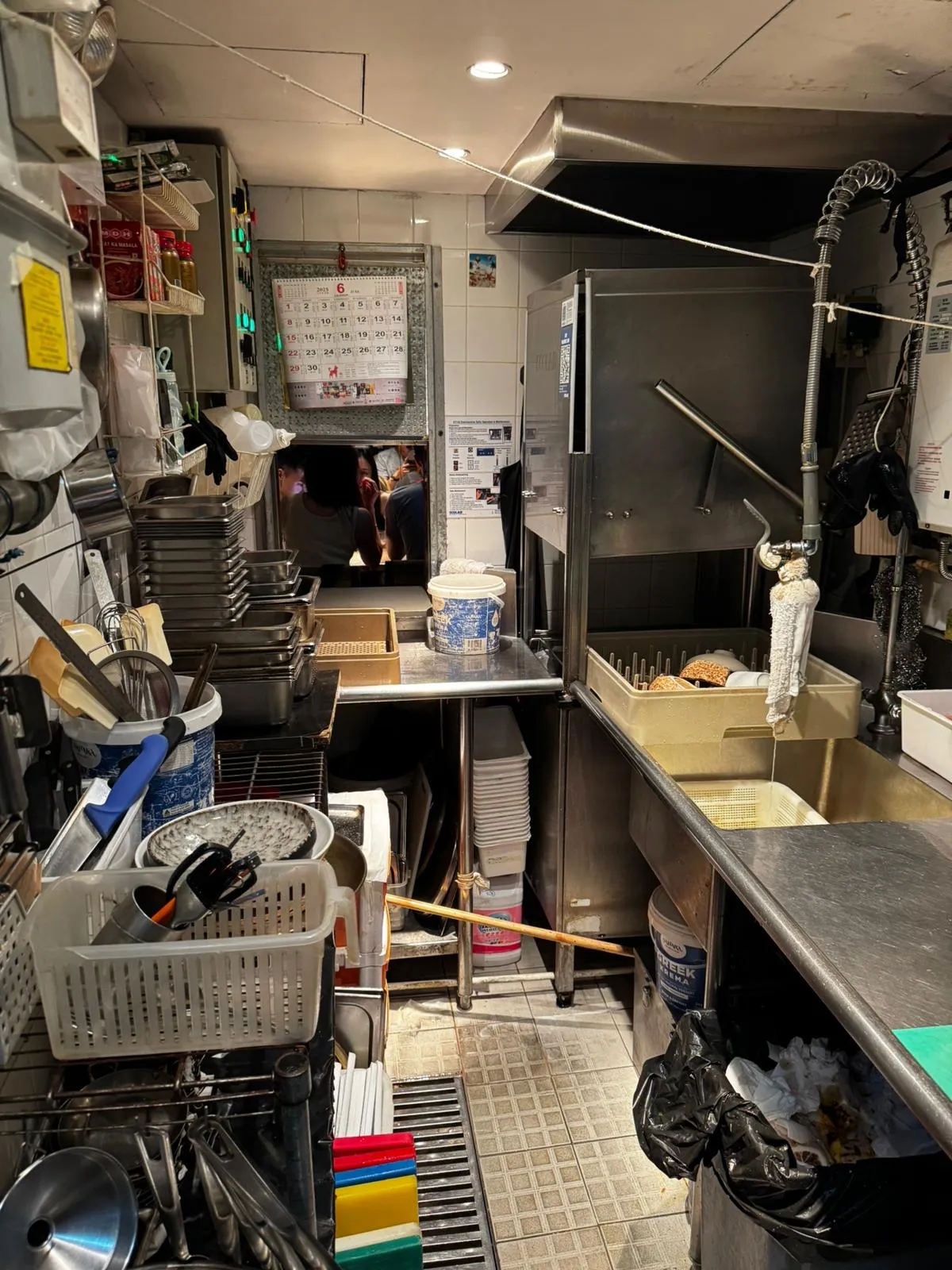 Ref#3654 is featured in a busy, compact restaurant kitchen filled with stacked dishes, utensils, and cleaning supplies. Stainless steel surfaces and a dishwashing area dominate, while people can be seen through a small back-wall window.