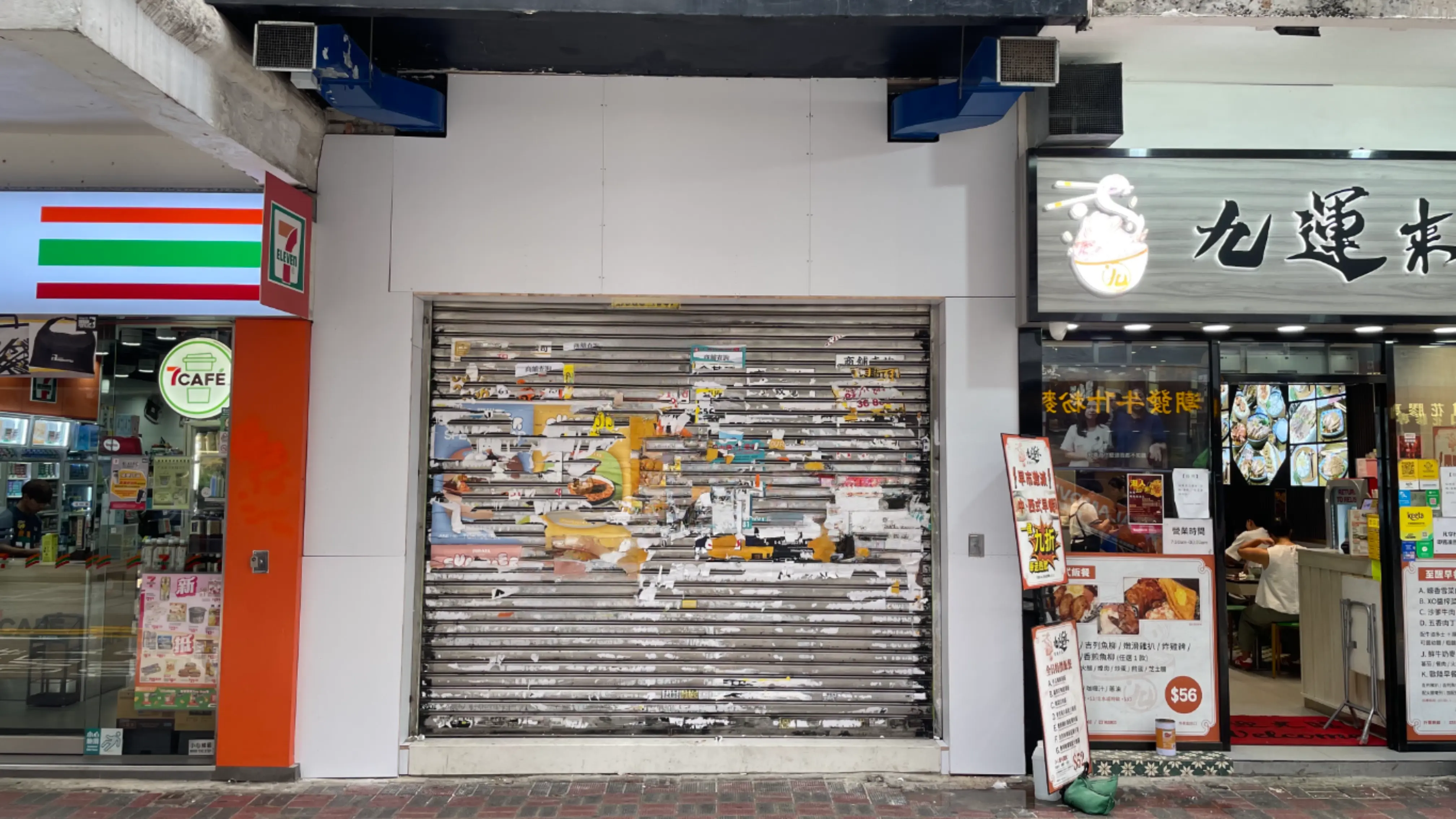 Ref#3653: A closed metal storefront shutter covered in torn, faded posters sits between a convenience store and a brightly-lit restaurant for rent, all along a bustling city sidewalk.