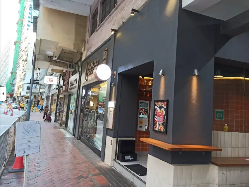 A city sidewalk featuring shops, a cafe with a dark facade, round sign, and vending machine at the entrance. A street sign and traffic cones sit on the red-brick walkway near shop rental opportunity Ref#3655.