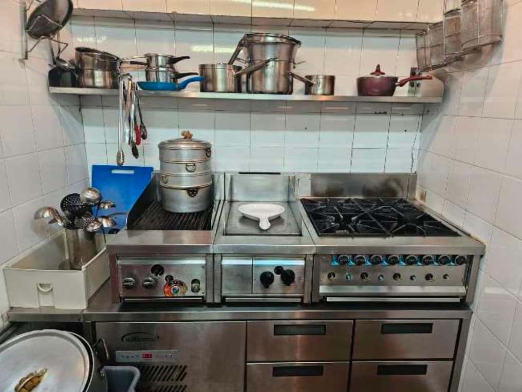 Ref#3632 is a commercial kitchen for rent, featuring metal pots and utensils on shelves, a multi-burner stovetop, grill, steamer, stainless steel drawers beneath, and white tiled walls in the background.