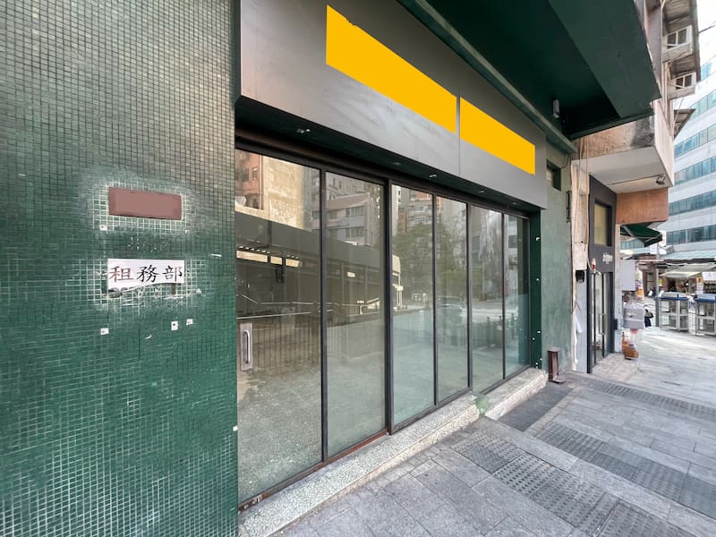 Ref#3639: A closed storefront with large glass windows and a dark green tiled exterior, perfect for shop rental. Two yellow rectangles cover signage above the entrance; Chinese characters appear on a sign to the left. Sidewalk visible in front.