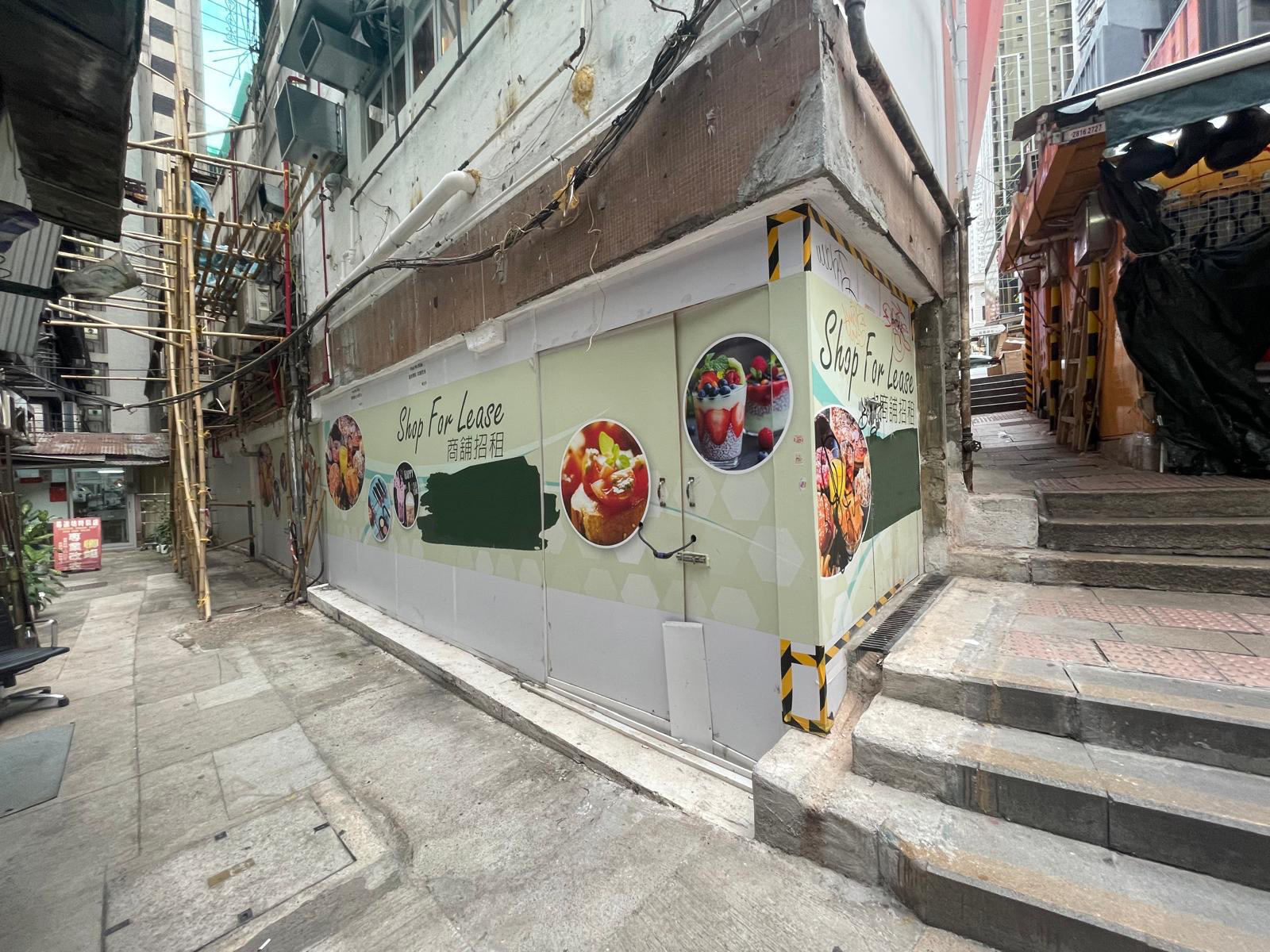 Ref#3628: Vacant corner restaurant storefront with shop rental signage and food images on the wall, situated among narrow alleyways with stairs ascending on the right.