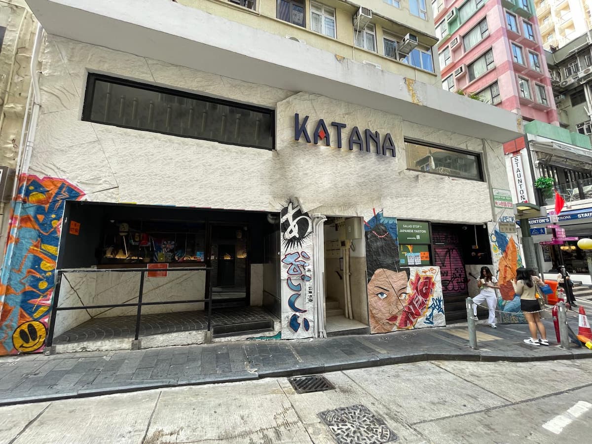Ref#3627: A building named KATANA features colorful murals with abstract faces and patterns on its exterior walls. Ideal for a shop rental, it's set against tall apartment buildings, with two people by the entrance on the sidewalk.