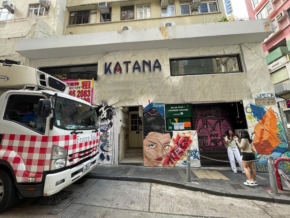 Street view of Ref#3627 with a KATANA sign, colorful murals, and a freezer truck out front. Two women take photos nearbya prime spot to rent shop or restaurant space in this lively urban area.