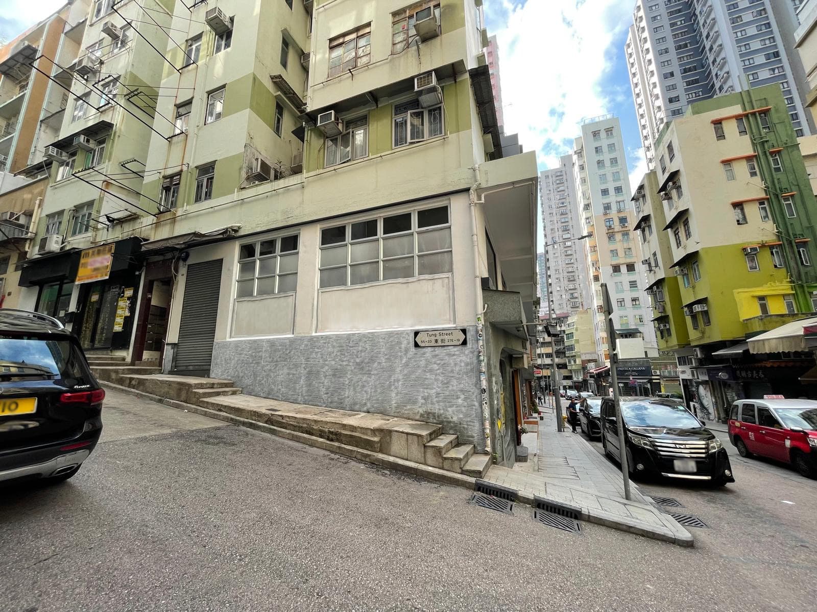 Ref#3629: Urban street with tall, closely packed apartments, some featuring balconies and air conditioners. Cars line both sides, and a small staircase leads to a corner building perfect for shop or restaurant rental.