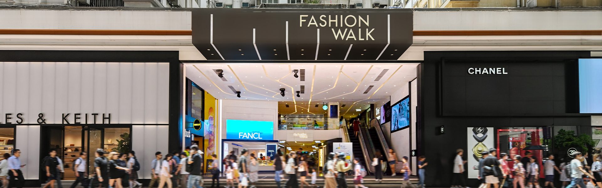 A lively street scene at Fashion Walk’s entrance (Ref#3621), showing glass doors, bright lights, people passing by stores, plus opportunities to rent shop or restaurant spaces.