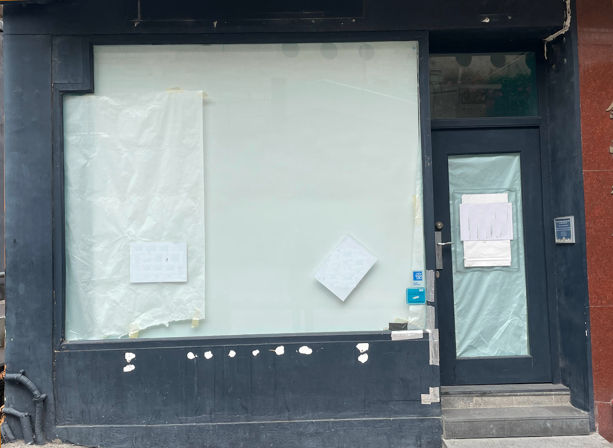 Ref#3619: Storefront with large windows and black exterior. Windows and door are covered with white paper and taped notices. Some white paint spots near bottom of facade. Ideal for rent as a shop or office space.
