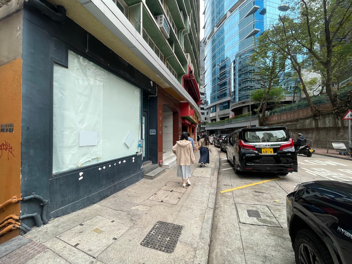 A woman walks down a narrow city sidewalk lined with parked cars and tall buildings, including Ref#3619—an office space with glass facades available for rent. Other pedestrians and trees line the street on the right.