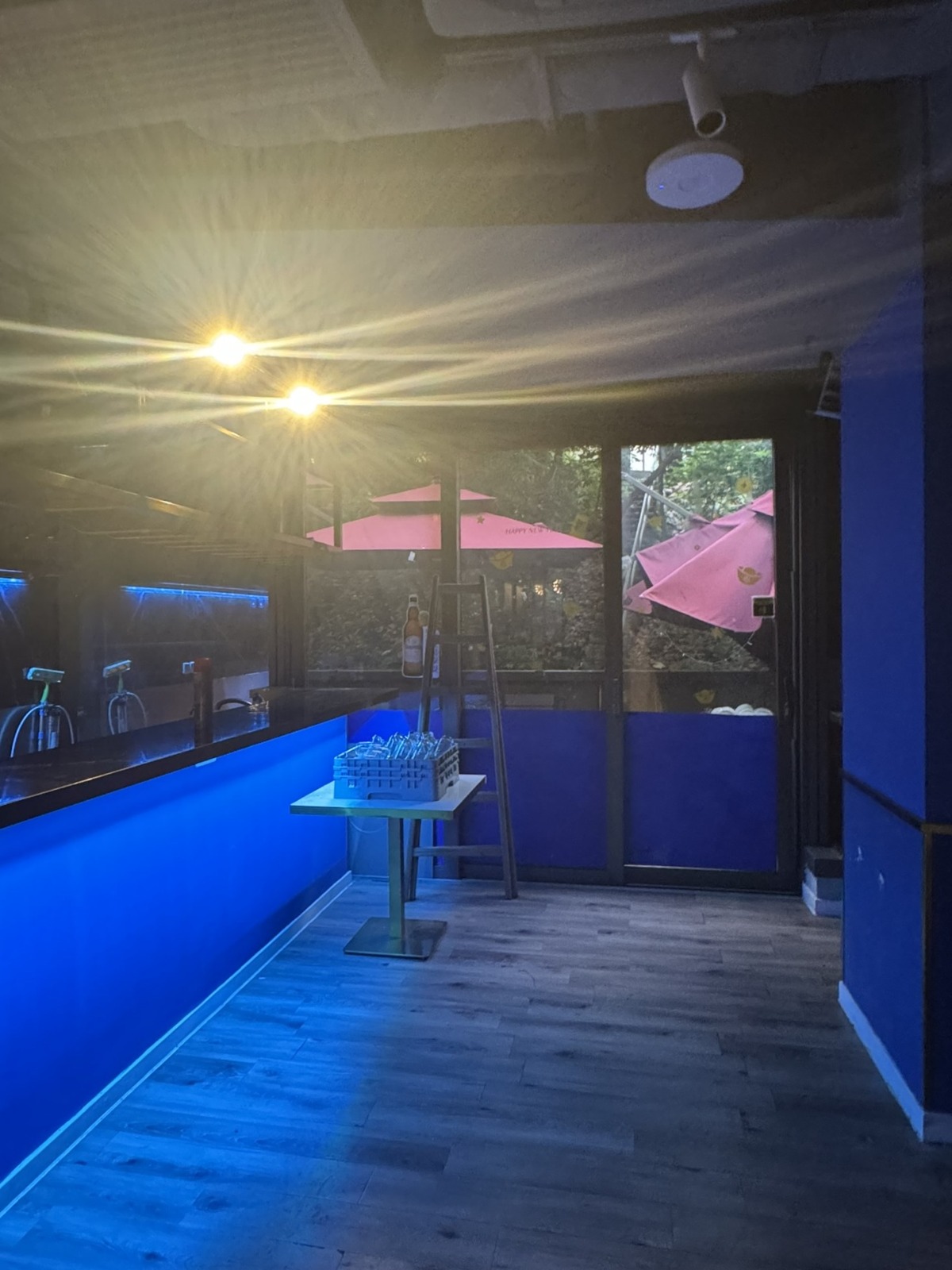 Ref#3613 features a dimly lit bar with blue walls and floors, stacked glasses on the counter, warm ceiling lights, and a window view of red umbrellas and greenery—an inviting space ideal to rent as a shop or restaurant.