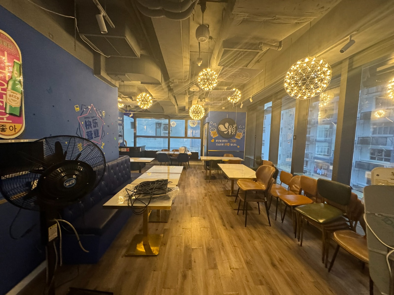 Ref#3613: Modern, unfurnished café with wooden floors, blue postered walls, hanging globe lights. Features tables and chairs by large windows for natural light; includes a standing fan and coiled cables on a table—ideal restaurant rental.