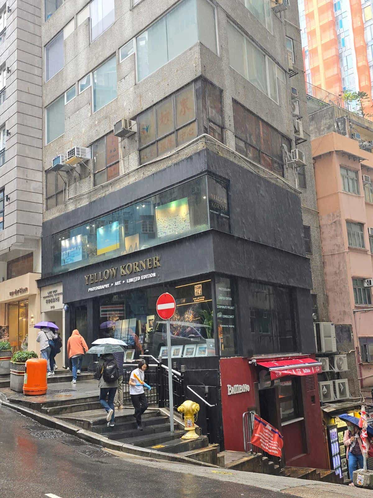 雨天下行人於中環街道上經過Yellow Korner，位於一座灰色多層建築物底層。