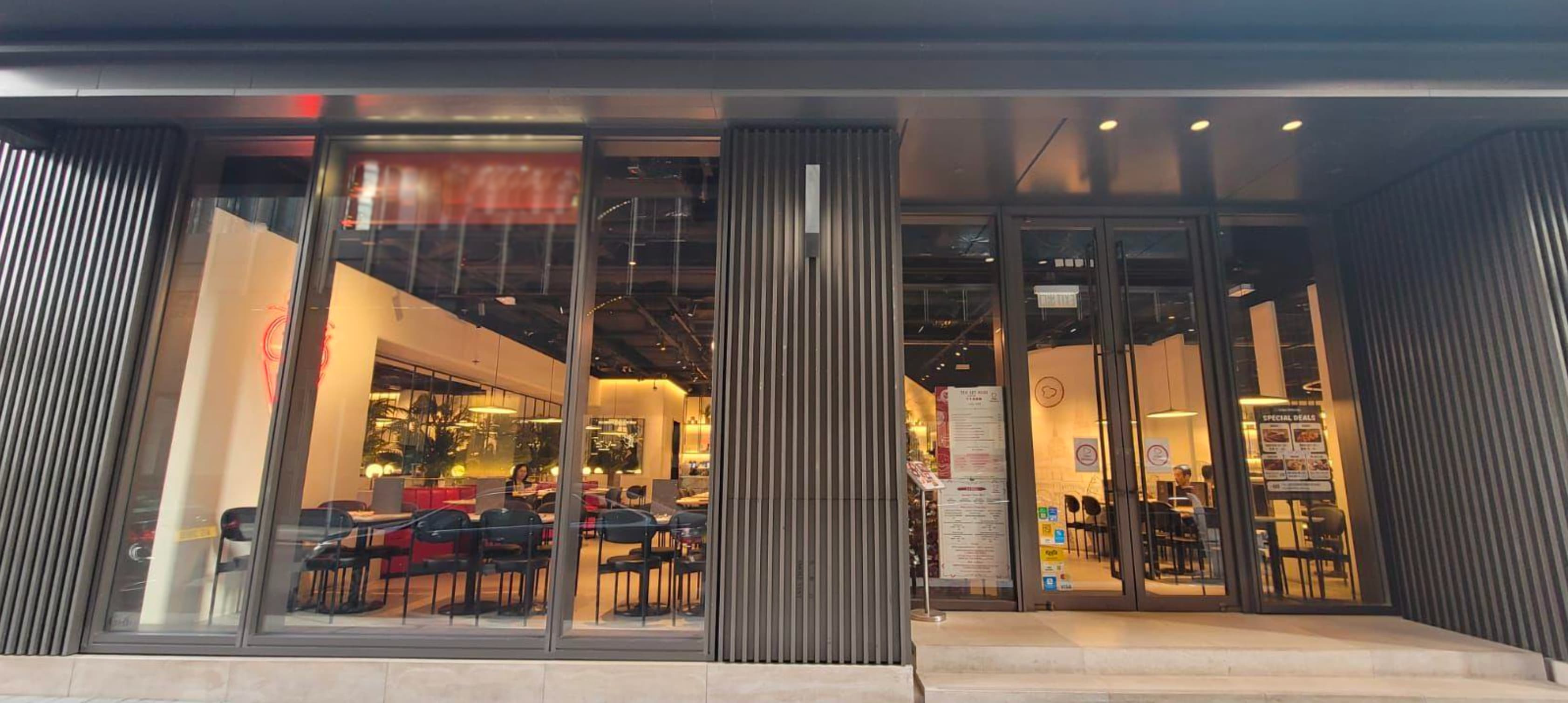 Street view of Ref#3611, a modern restaurant rental featuring large floor-to-ceiling windows, black vertical panels, and a warmly lit interior with tables, chairs, and minimalistic decor.