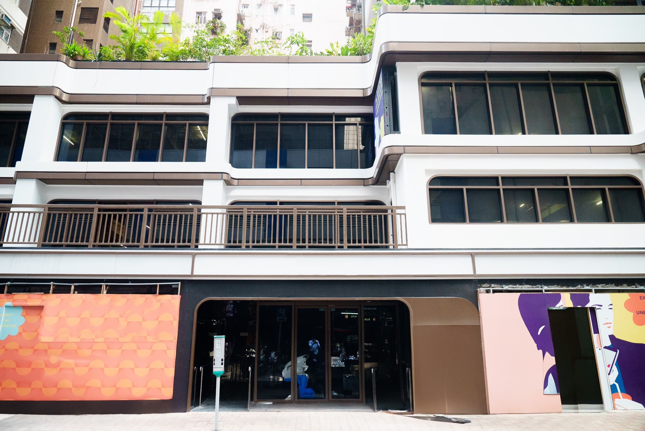 Ref#3610: Modern building with large windows, white and brown geometric facade, colorful ground-floor muralsideal for restaurant or shop rental. Rooftop greenery and a sidewalk parking meter complete the features.