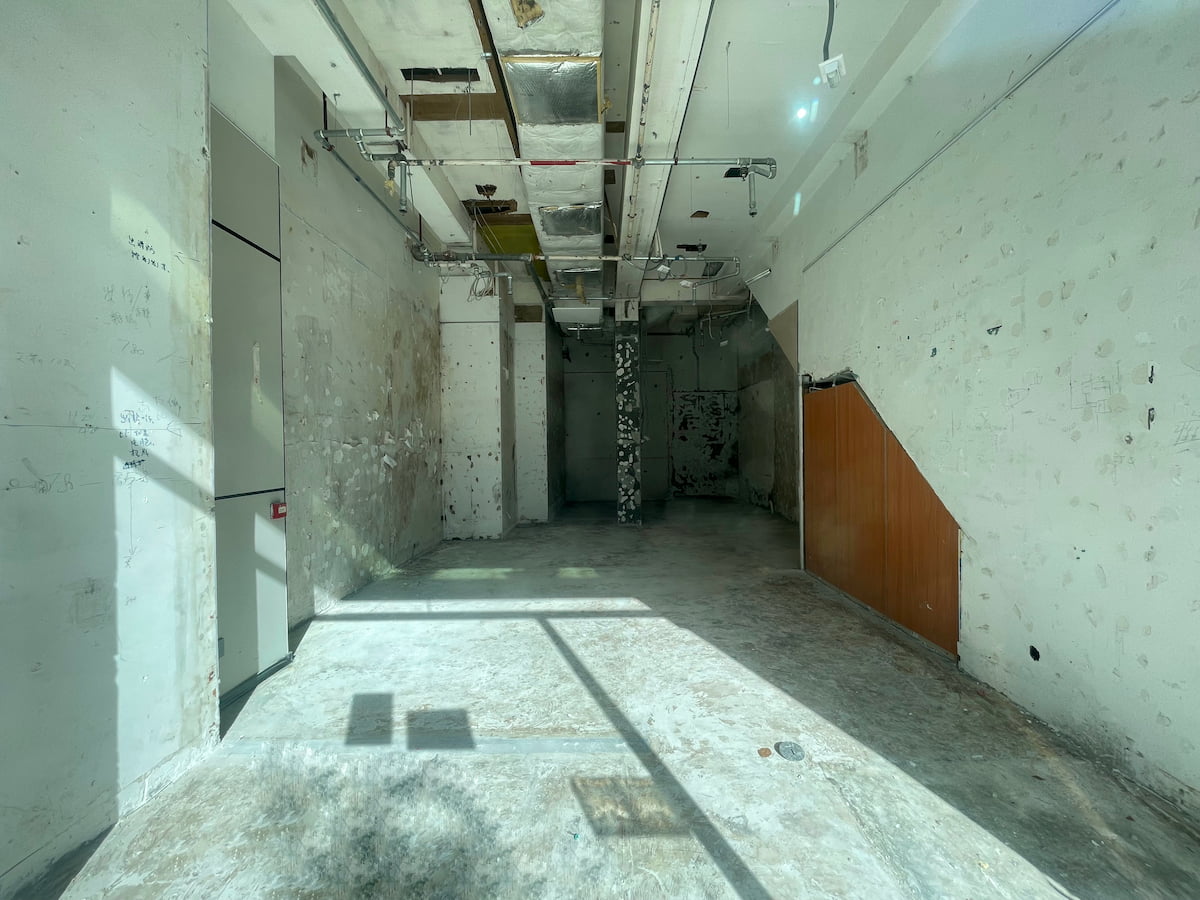 Ref#3604: Sunlit room with bare concrete floors, patched white walls, exposed ceiling pipes, and a small area of brown paneling under the staircase10ideal for rent as a shop or office.