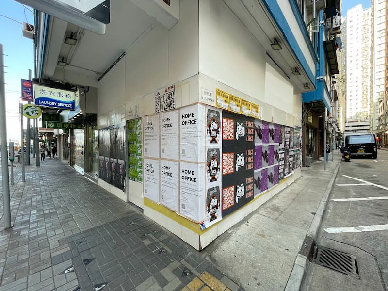 On an urban street corner, posters cover walls
	66including some for Ref#3607	64while a laundromat sign hangs above the sidewalk and tall buildings rise on both sides.